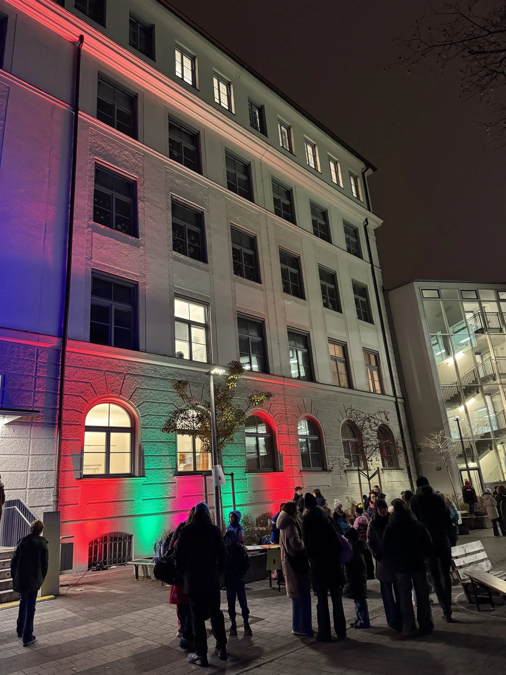 Die Schulfassade am Abend in Rot, Grün und Pink angestrahlt, davor Besucherinnen und Besucher
