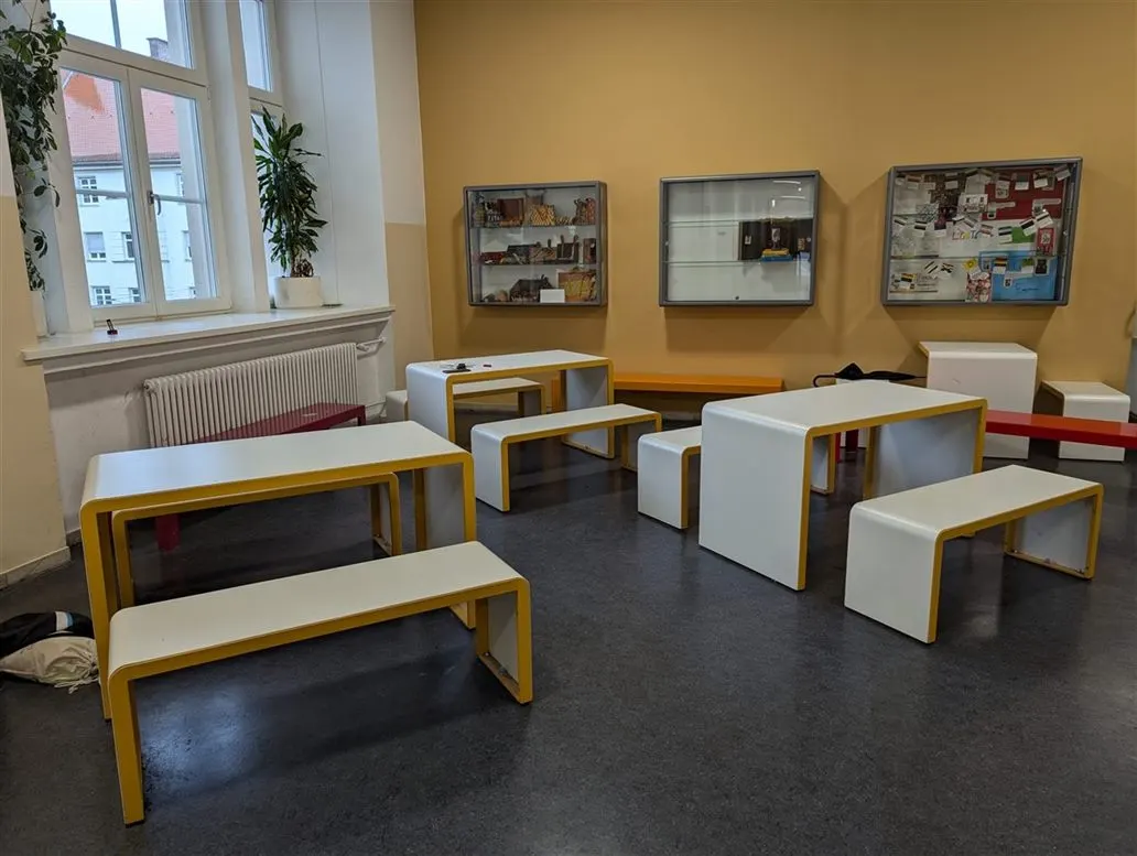 Sitzgruppen aus weißen Tischen und Bänken vor einer gelben Wand im Foyer des MTG
