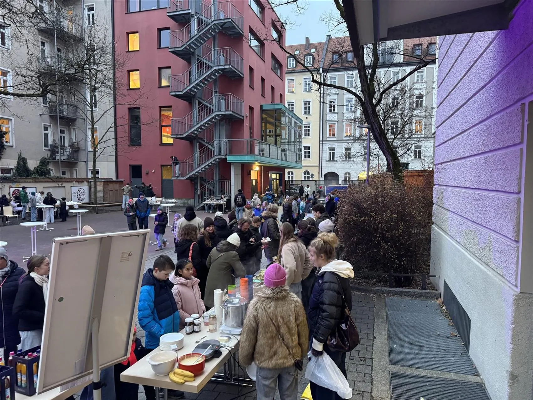 Winterabend im Pausenhof: Stehtische, ein Crêpes-Stand und viele Gäste in Wintermänteln vor dem beleuchteten Schulhaus