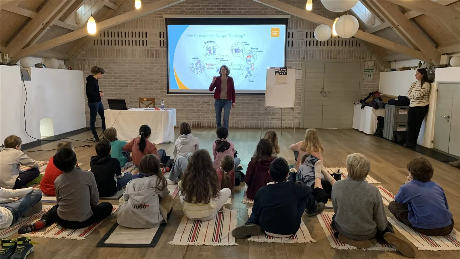 Eine Klasse sitzt im Kreis auf dem Boden vor einer Präsentation zum Thema Design Thinking