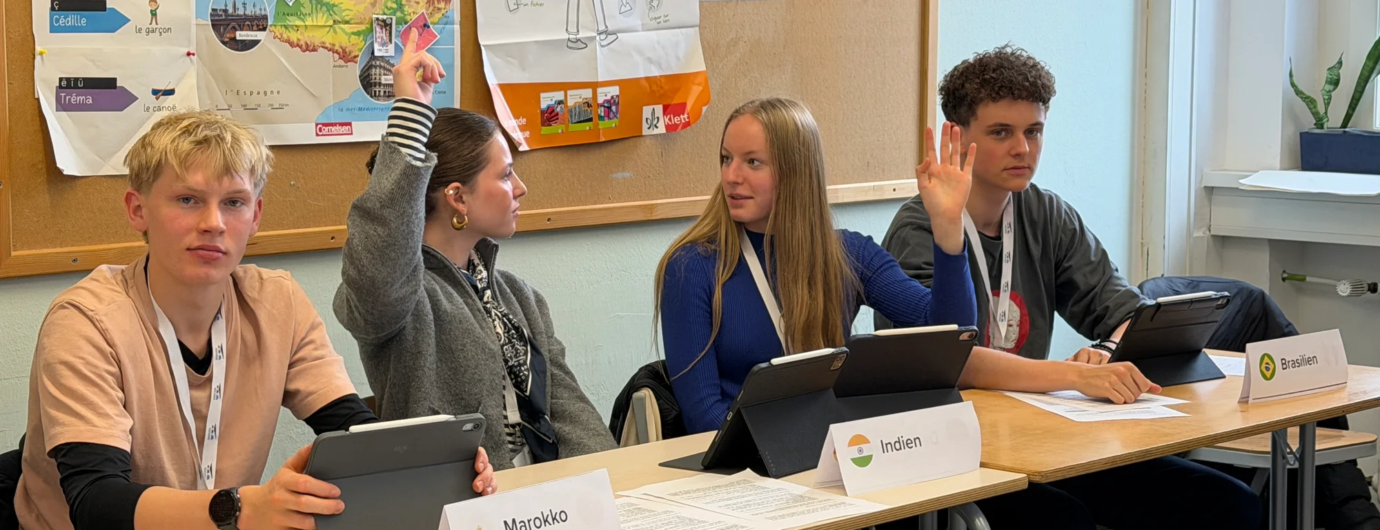 Schülerinnen und Schüler an Tischen mit Ländernamen bei einem Planspiel, eine Hand ist erhoben