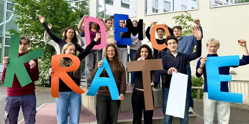 Schülerinnen und Schüler halten Buchstaben hoch, die das Wort DEMOKRATIE ergeben