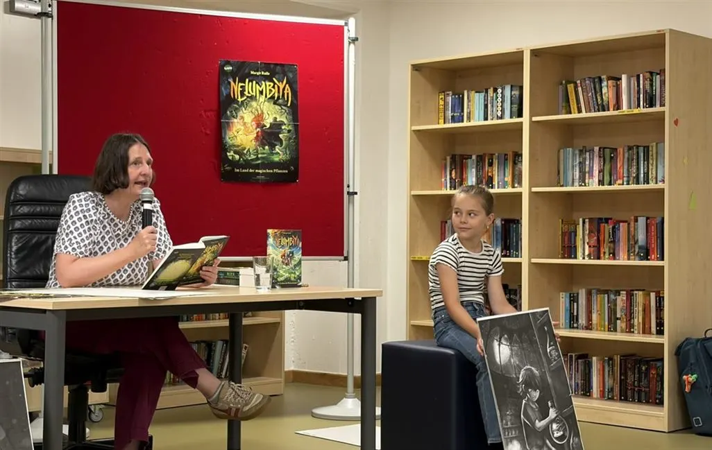 Die Autorin Margit Ruile liest in der Schulbibliothek aus ihrem Buch, daneben sitzt eine Schülerin mit einer Illustration