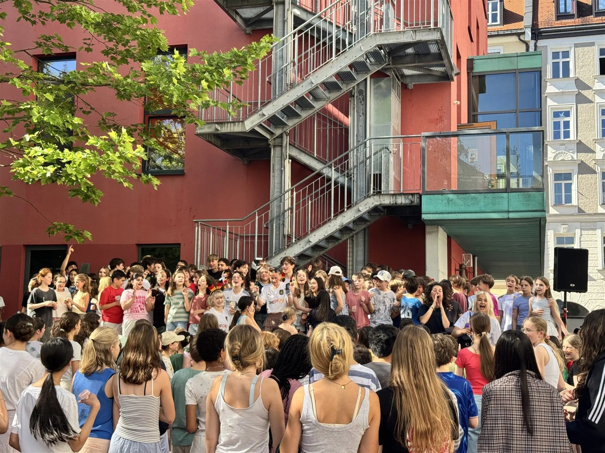 Schülerinnen und Schüler singen und jubeln beim Sommerfest im Innenhof, dahinter der rote Neubau